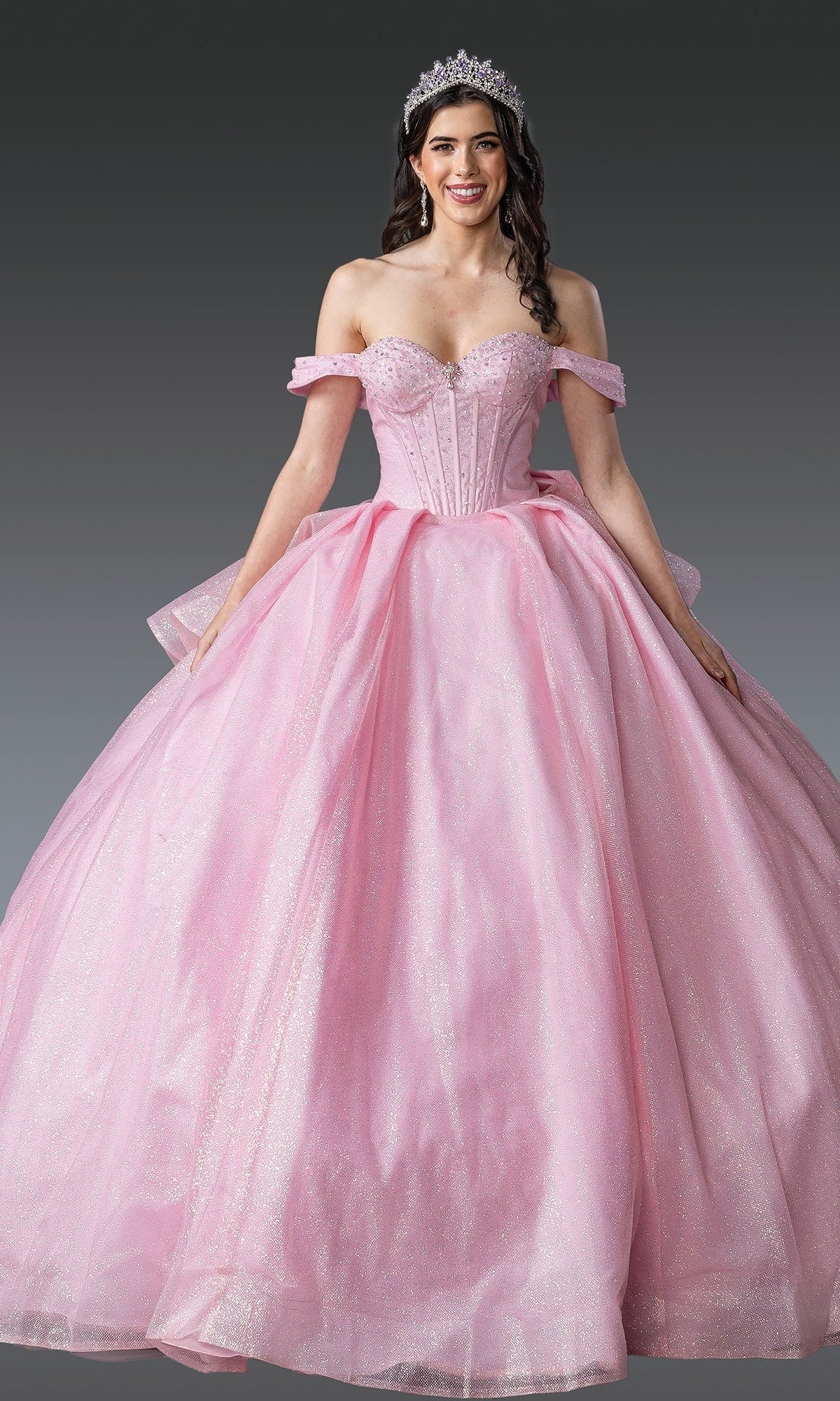 A woman wears the Quinceanera Dress 1937 by Dancing Queen, a sparkling pink off-the-shoulder ball gown, paired with a tiara, against a plain gray background.